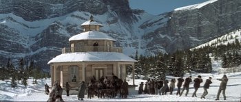 Movie still from “The Claim” (2000), directed by Michael Winterbottom – A group of people standing around a building in the snow; Extreme Wide shot, High angle