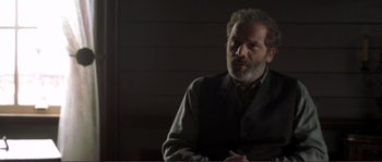 Movie still from “The Claim” (2000), directed by Michael Winterbottom – A man with a gray beard is sitting in a dark room; Medium shot, Over the shoulder angle