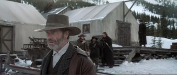 Movie still from “The Claim” (2000), directed by Michael Winterbottom – A group of people standing in the snow near a building; Medium shot, Over the shoulder angle