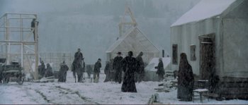 Movie still from “The Claim” (2000), directed by Michael Winterbottom – A group of people standing in the snow near a building; Extreme Wide shot, High angle