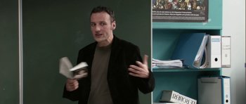 Movie still from “The Class” (2008), directed by Laurent Cantet – A man in a black jacket is holding a book and gesturing with his hands; Medium shot, Over the shoulder angle