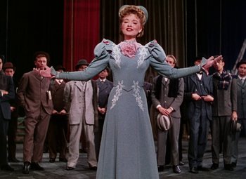 Movie still from “The Climax” (1944), directed by George Waggner – A woman wearing a dress and hat in front of a group of men; Medium shot, Low angle