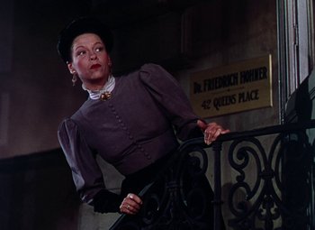 Movie still from “The Climax” (1944), directed by George Waggner – A woman wearing a hat and a skirt standing next to a railing in front of a building; Medium shot, Low angle