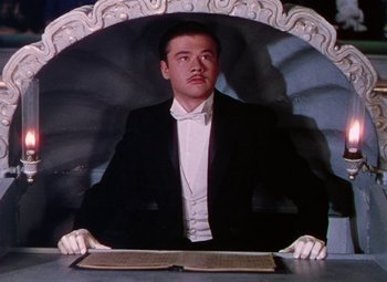 Movie still from “The Climax” (1944), directed by George Waggner – A man in a suit sitting at a table with an open book in front of him; Medium shot, Low angle