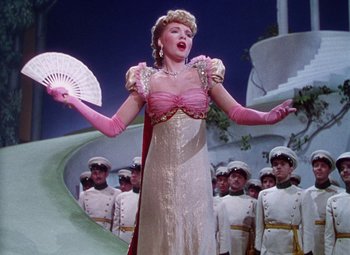 Movie still from “The Climax” (1944), directed by George Waggner – A woman in a pink dress holding a fan in front of a group of men; Medium shot, Low angle
