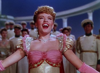 Movie still from “The Climax” (1944), directed by George Waggner – A woman in a pink and gold dress singing in front of a group of people; Medium shot, Low angle