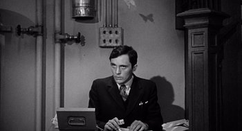 Movie still from “The Collector” (1965), directed by William Wyler – A black and white photo of a man sitting at a desk with a computer; Medium shot, High angle