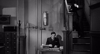 Movie still from “The Collector” (1965), directed by William Wyler – A black and white photo of a man sitting at a desk; Medium shot, High angle