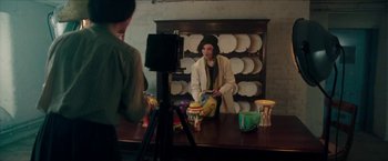 Movie still from “The Colour Room” (2021), directed by Claire McCarthy – A man sitting in front of a camera on top of a wooden table; Medium shot, Over the shoulder angle