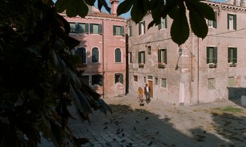Movie still from “The Comfort of Strangers” (1990), directed by Paul Schrader – A couple of people walking down a street next to a building; Extreme Wide shot, High angle