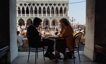 Movie still from “The Comfort of Strangers” (1990), directed by Paul Schrader – A man and a woman sitting at a table in front of a building; Wide shot, Over the shoulder angle