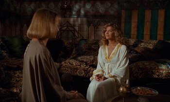 Movie still from “The Comfort of Strangers” (1990), directed by Paul Schrader – A woman in a white robe sitting on a couch talking to another woman; Medium shot, Over the shoulder angle