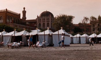 Movie still from “The Comfort of Strangers” (1990), directed by Paul Schrader – A group of people standing next to tents on a beach; Extreme Wide shot, High angle