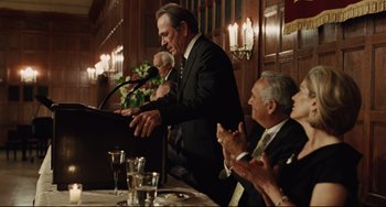 Movie still from “The Company Men” (2010), directed by John Wells – A man standing at a podium in front of a microphone; Medium shot, Over the shoulder angle