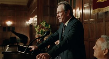 Movie still from “The Company Men” (2010), directed by John Wells – An older man in a suit is speaking into a microphone; Medium shot, Over the shoulder angle