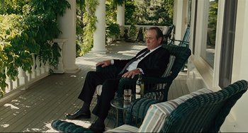 Movie still from “The Company Men” (2010), directed by John Wells – A man in a suit and tie sitting in a chair on a porch; Medium shot, Over the shoulder angle