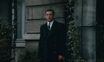 Movie still from “The Confession” (1970), directed by Costa-Gavras – A man in a suit and tie standing in front of a building; Medium shot, Over the shoulder angle