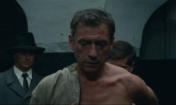 Movie still from “The Confession” (1970), directed by Costa-Gavras – A man with no shirt is standing next to another man; Close Up shot, Low angle