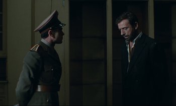 Movie still from “The Confession” (1970), directed by Costa-Gavras – A man in a suit and a man in a military uniform stand next to each other in front of a door; Medium shot, Low angle