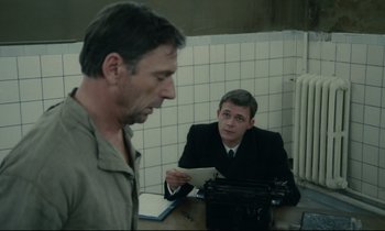 Movie still from “The Confession” (1970), directed by Costa-Gavras – Two men are sitting at a table and one of the men is holding papers; Medium shot, Over the shoulder angle