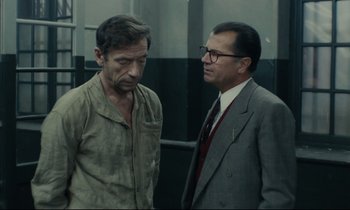 Movie still from “The Confession” (1970), directed by Costa-Gavras – A man and a man in a suit talking to each other; Medium shot, Over the shoulder angle