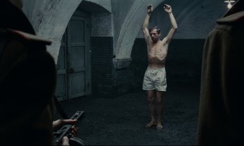 Movie still from “The Confession” (1970), directed by Costa-Gavras – A man standing in the middle of a room with his arms raised; Wide shot, Low angle
