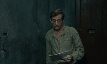 Movie still from “The Confession” (1970), directed by Costa-Gavras – A man holding a book while standing in front of a wall; Medium shot, High angle