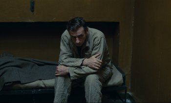 Movie still from “The Confession” (1970), directed by Costa-Gavras – A man sitting on top of a bed in a room; Medium shot, High angle