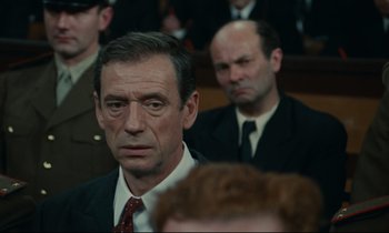 Movie still from “The Confession” (1970), directed by Costa-Gavras – A man in a suit and tie in front of other men in suits; Close Up shot, Over the shoulder angle