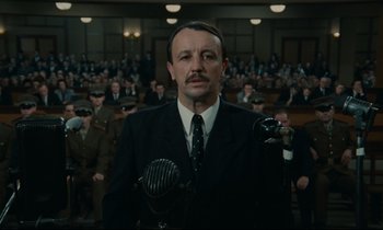 Movie still from “The Confession” (1970), directed by Costa-Gavras – A man in a suit and tie standing in front of an audience; Medium shot, Low angle