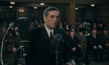 Movie still from “The Confession” (1970), directed by Costa-Gavras – An older man wearing a suit and tie is standing in front of a microphone in front of an audience; Medium shot, Low angle