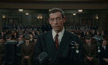 Movie still from “The Confession” (1970), directed by Costa-Gavras – A man in a suit and tie is speaking into a microphone; Medium shot, Low angle