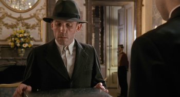 Movie still from “The Counterfeiters” (2007), directed by Stefan Ruzowitzky – A man in a suit and a hat is standing in front of a table; Medium shot, Over the shoulder angle