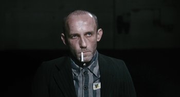Movie still from “The Counterfeiters” (2007), directed by Stefan Ruzowitzky – A man in a suit smoking a cigarette in the dark; Medium shot, Over the shoulder angle