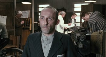 Movie still from “The Counterfeiters” (2007), directed by Stefan Ruzowitzky – A man in a suit and tie standing in front of a machine; Medium shot, Low angle