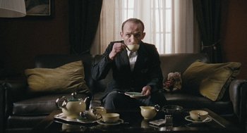 Movie still from “The Counterfeiters” (2007), directed by Stefan Ruzowitzky – A man sitting on top of a black couch eating; Medium shot, Low angle