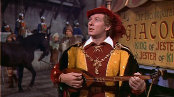 Movie still from “The Court Jester” (1955), directed by Melvin Frank – A man dressed in medieval clothing holding a guitar in front of a group of people; Medium shot, Low angle