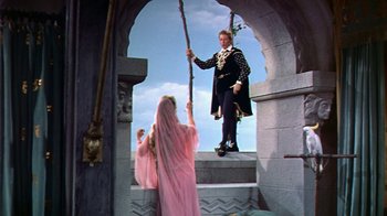 Movie still from “The Court Jester” (1955), directed by Melvin Frank – A woman in a pink dress stands on steps as a man in a black suit holds a stick; Wide shot, Low angle
