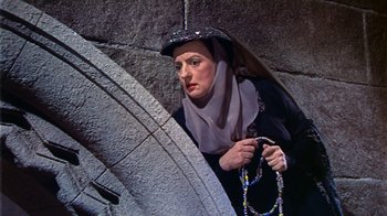 Movie still from “The Court Jester” (1955), directed by Melvin Frank – A woman in a black dress and a hat holding a necklace; Medium shot, Low angle