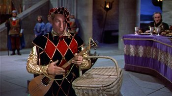 Movie still from “The Court Jester” (1955), directed by Melvin Frank – A man dressed as a harlequin holding a guitar and standing next to a wicker basket; Medium shot, High angle