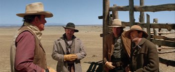 Movie still from “The Cowboys” (1972), directed by Mark Rydell – A couple of men standing next to each other on top of a dirt field; Medium shot, Over the shoulder angle