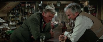 Movie still from “The Cowboys” (1972), directed by Mark Rydell – Two men sitting at a bar with a drink in front of them; Medium shot, Over the shoulder angle