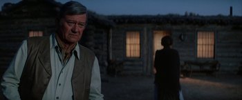 Movie still from “The Cowboys” (1972), directed by Mark Rydell – An older man standing in front of a log cabin; Medium shot, Over the shoulder angle
