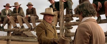 Movie still from “The Cowboys” (1972), directed by Mark Rydell – An old man wearing a hat sitting next to a wooden fence; Medium shot, Over the shoulder angle