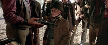 Movie still from “The Cowboys” (1972), directed by Mark Rydell – A young boy in a brown jacket holding a gun; Medium shot, Over the shoulder angle