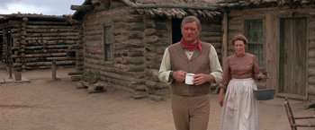 Movie still from “The Cowboys” (1972), directed by Mark Rydell – A man holding a cup while standing in front of a log cabin; Medium shot, Over the shoulder angle