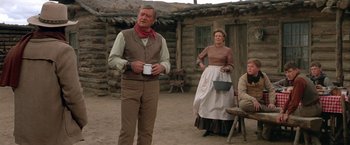 Movie still from “The Cowboys” (1972), directed by Mark Rydell – A man and a woman standing next to each other in front of a log cabin; Wide shot, Over the shoulder angle