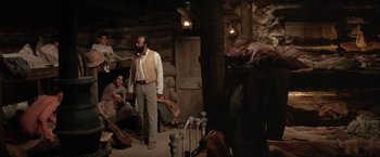 Movie still from “The Cowboys” (1972), directed by Mark Rydell – A man standing in front of a group of people in a room; Wide shot, High angle