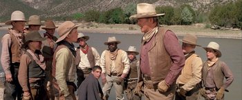 Movie still from “The Cowboys” (1972), directed by Mark Rydell – A group of men standing next to a body of water; Medium shot, Over the shoulder angle