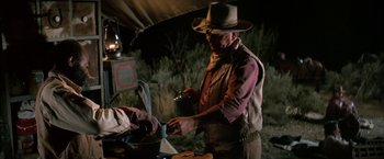 Movie still from “The Cowboys” (1972), directed by Mark Rydell – A man in a cowboy hat cooking food in an outdoor kitchen; Medium shot, Over the shoulder angle